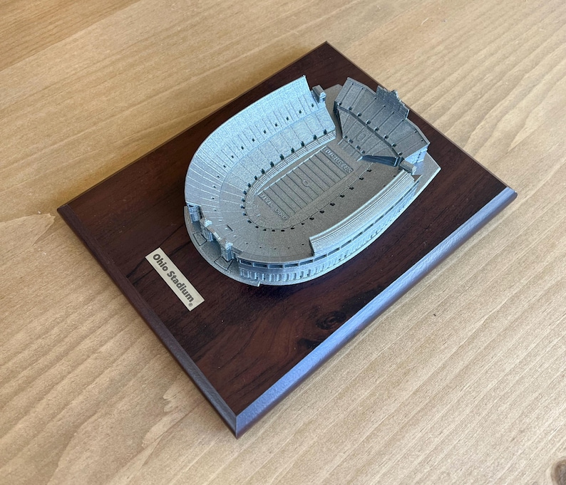 Może przedstawiać: Srebrny model stadionu Ohio na ciemnym drewnianym cokole. Na cokole znajduje się tabliczka z napisem "Ohio Stadium."