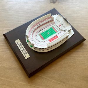Może przedstawiać: Miniaturowa replika stadionu Ohio, domu Ohio State Buckeyes, na ciemnobrązowej drewnianej podstawie. Stadion jest biały z czerwonymi siedzeniami i zielonym boiskiem. Tekst "Ohio Stadium Ohio State Buckeyes" znajduje się na złotej tabliczce.