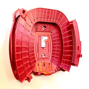 May include: Red plastic light switch cover shaped like a football stadium. The switch cover has a white light switch and the text "GEORGIA" is visible on the bottom.