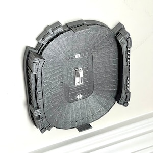 May include: A 3D printed light switch cover shaped like a football stadium. The switch cover is gray and has a white light switch in the center. The stadium is a detailed replica of a real stadium.