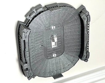 May include: A 3D printed light switch cover shaped like a football stadium. The switch cover is gray and has a white light switch in the center. The stadium is a detailed replica of a real stadium.