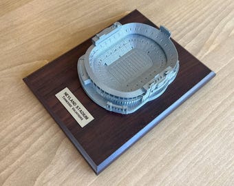 May include: A 3D model of Neyland Stadium, home of the Tennessee Volunteers, on a dark wood base. The model is made of a silver-colored material and shows the stadium's seating, field, and surrounding area.