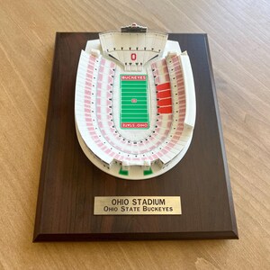 Może przedstawiać: Biały plastikowy model stadionu Ohio, domu Ohio State Buckeyes, na ciemnobrązowej drewnianej tabliczce. Tabliczka ma złotą tabliczkę z tekstem "OHIO STADIUM OHIO STATE BUCKEYES".