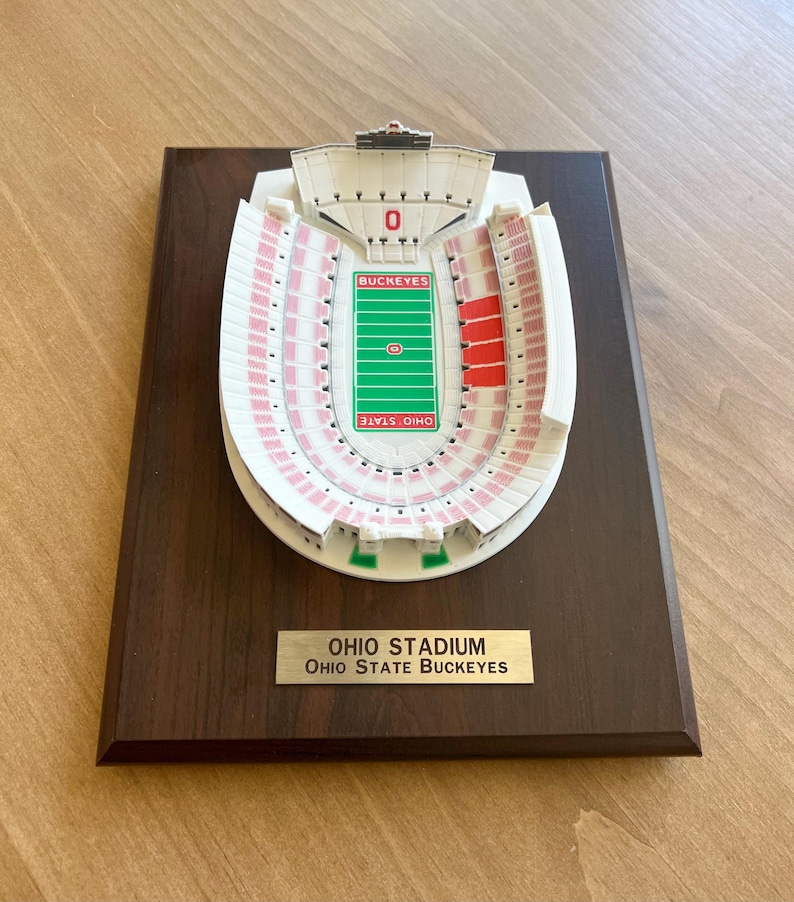 Może przedstawiać: Biały plastikowy model stadionu Ohio, domu Ohio State Buckeyes, na ciemnobrązowej drewnianej tabliczce. Tabliczka ma złotą tabliczkę z tekstem "OHIO STADIUM OHIO STATE BUCKEYES".