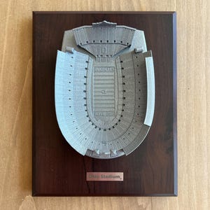 Może przedstawiać: Srebrny model 3D Ohio Stadium, dużego stadionu piłkarskiego, na ciemnobrązowej drewnianej tabliczce. Tabliczka zawiera napis "Ohio Stadium".