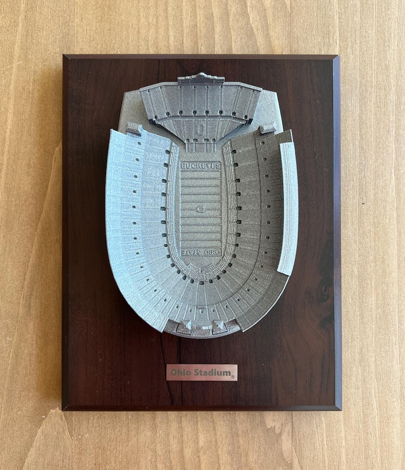 Może przedstawiać: Srebrny model 3D Ohio Stadium, dużego stadionu piłkarskiego, na ciemnobrązowej drewnianej tabliczce. Tabliczka zawiera napis "Ohio Stadium".