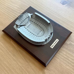 Może przedstawiać: Miniaturowa replika stadionu Ohio Stadium, dużego, owalnego stadionu z brązową drewnianą podstawą. Stadion jest wykonany z metalu i ma szare wykończenie. Podstawa ma małą tabliczkę z napisem "Ohio Stadium."