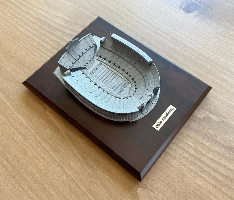 Może przedstawiać: Miniaturowa replika stadionu Ohio Stadium, dużego, owalnego stadionu z brązową drewnianą podstawą. Stadion jest wykonany z metalu i ma szare wykończenie. Podstawa ma małą tabliczkę z napisem "Ohio Stadium."