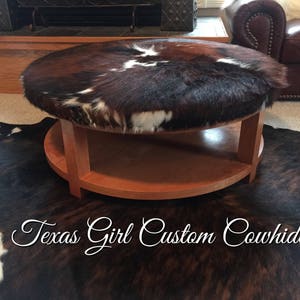 May include: A round, wooden coffee table with a brown and white cowhide top. The table has a lower shelf and is sitting on a brown and white cowhide rug. The text "Texas Girl Custom Cowhide" is visible in the image.