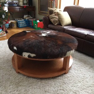 May include: A round wooden coffee table with a brown and white cowhide top. The table has a lower shelf and is sitting on a white shag rug.