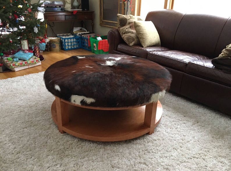 May include: A round wooden coffee table with a brown and white cowhide top. The table has a lower shelf and is sitting on a white shag rug.