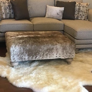 May include: A white faux fur rug with a brown and white cowhide ottoman in front of a beige sectional sofa with pillows.