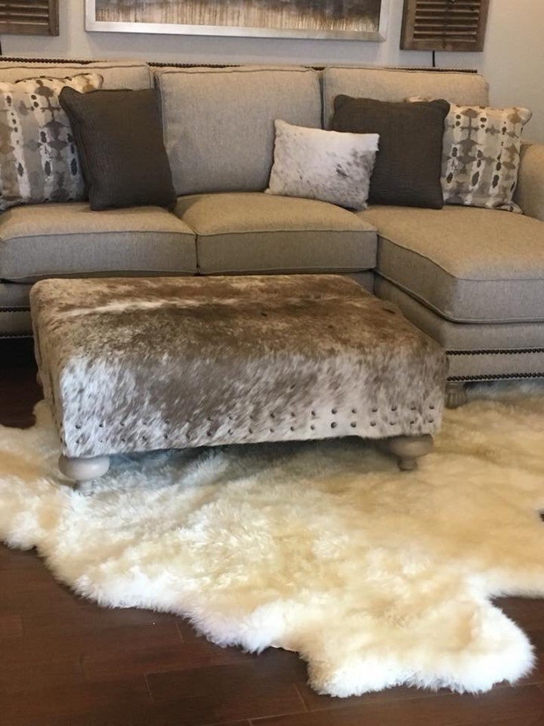May include: A white faux fur rug with a brown and white cowhide ottoman in front of a beige sectional sofa with pillows.