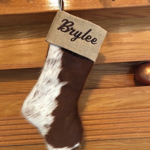 May include: A brown and white cowhide Christmas stocking with a burlap cuff. The cuff is embroidered with the name "Brylee".