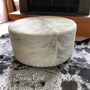 May include: A round, white cowhide ottoman with silver studs around the bottom edge. The ottoman is sitting on a black and white cowhide rug. The text "Texas Girl Custom Cowhide" is written on the rug.