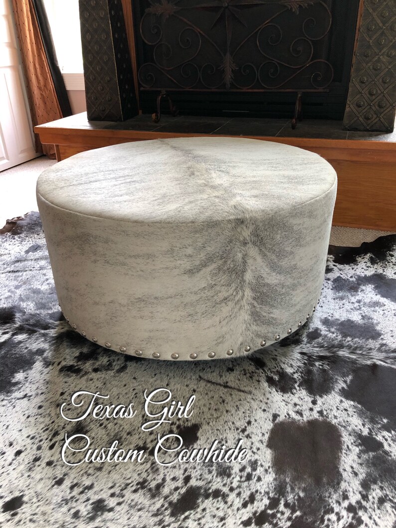 May include: A round, white cowhide ottoman with silver studs around the bottom edge. The ottoman is sitting on a black and white cowhide rug. The text "Texas Girl Custom Cowhide" is written on the rug.