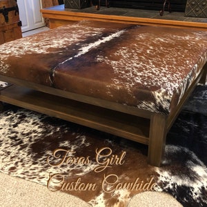 May include: A brown wooden coffee table with a cowhide top and a lower shelf. The table is sitting on a cowhide rug.