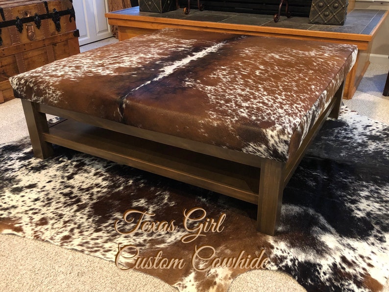 May include: A brown wooden coffee table with a cowhide top and a lower shelf. The table is sitting on a cowhide rug.