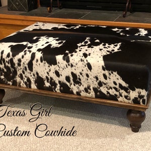 May include: A black and white cowhide ottoman with a wooden frame and four turned legs. The ottoman is decorated with silver studs. The text "Texas Girl Custom Cowhide" is visible in the image.