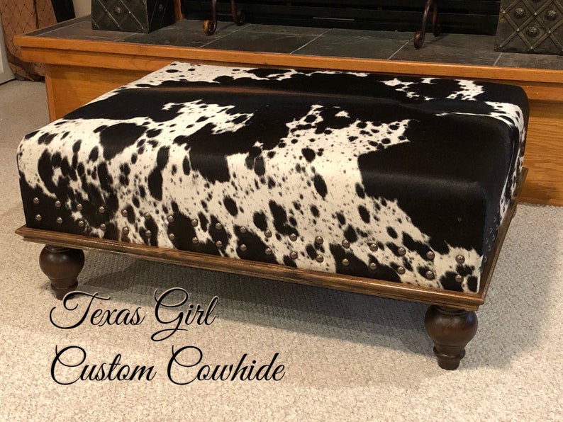 May include: A black and white cowhide ottoman with a wooden frame and four turned legs. The ottoman is decorated with silver studs. The text "Texas Girl Custom Cowhide" is visible in the image.