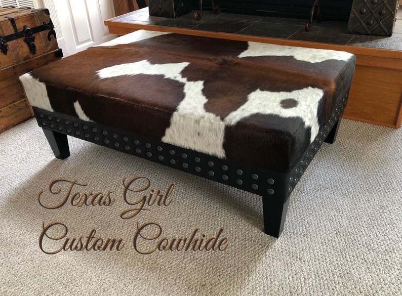 May include: A brown and white cowhide ottoman with black legs and silver studs around the edge. The ottoman is on a beige carpet. The text "Texas Girl Custom Cowhide" is written on the carpet in front of the ottoman.