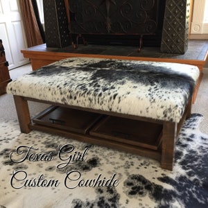 May include: A brown wooden coffee table with a black and white cowhide top. The table has two pull-out drawers and is sitting on a black and white cowhide rug. The text "Texas Girl Custom Cowhide" is written on the rug.