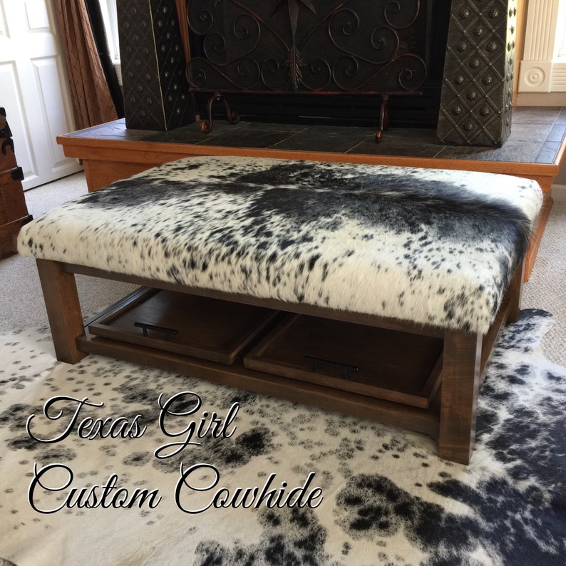 May include: A brown wooden coffee table with a black and white cowhide top. The table has two pull-out drawers and is sitting on a black and white cowhide rug. The text "Texas Girl Custom Cowhide" is written on the rug.