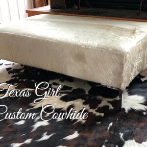 May include: White cowhide ottoman with chrome legs on a brown and white cowhide rug. The rug has the text "Texas Girl Custom Cowhide" on it.
