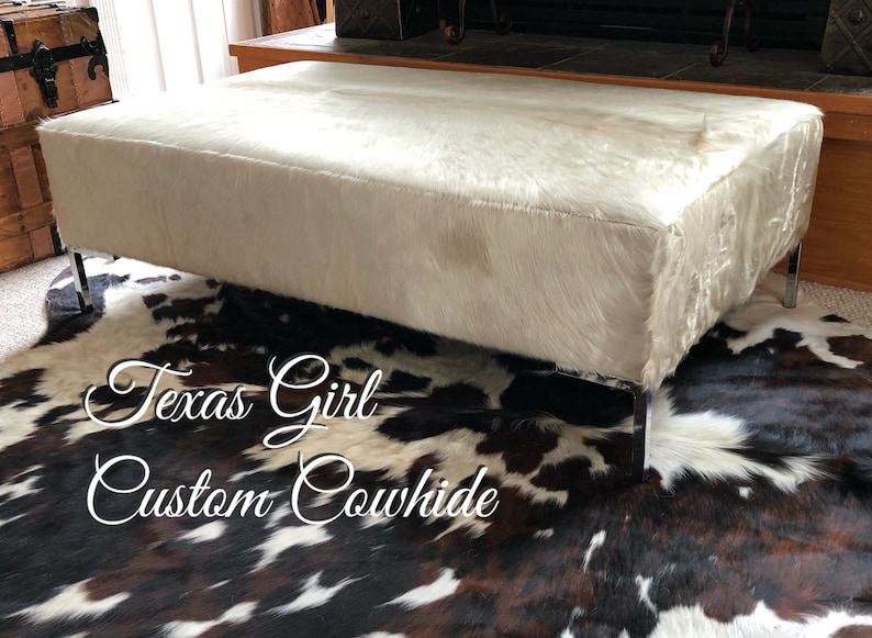 May include: White cowhide ottoman with chrome legs on a brown and white cowhide rug. The rug has the text "Texas Girl Custom Cowhide" on it.