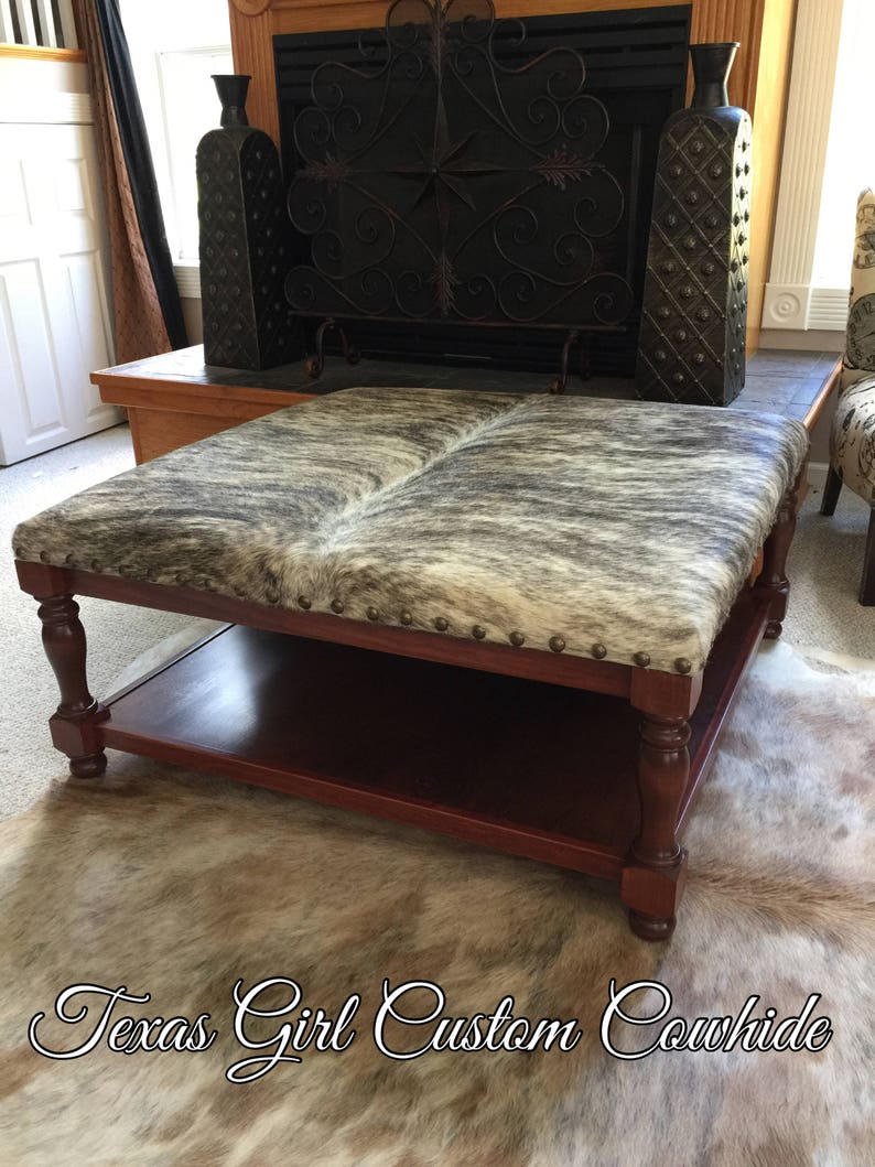 May include: A brown wooden coffee table with a cowhide top and a lower shelf. The table is sitting on a cowhide rug. The text "Texas Girl Custom Cowhide" is visible in the bottom right corner of the image.