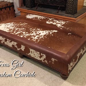 May include: A large, rectangular ottoman upholstered in brown leather with a cowhide top. The cowhide is brown and white with a spotted pattern. The ottoman has four wooden legs.