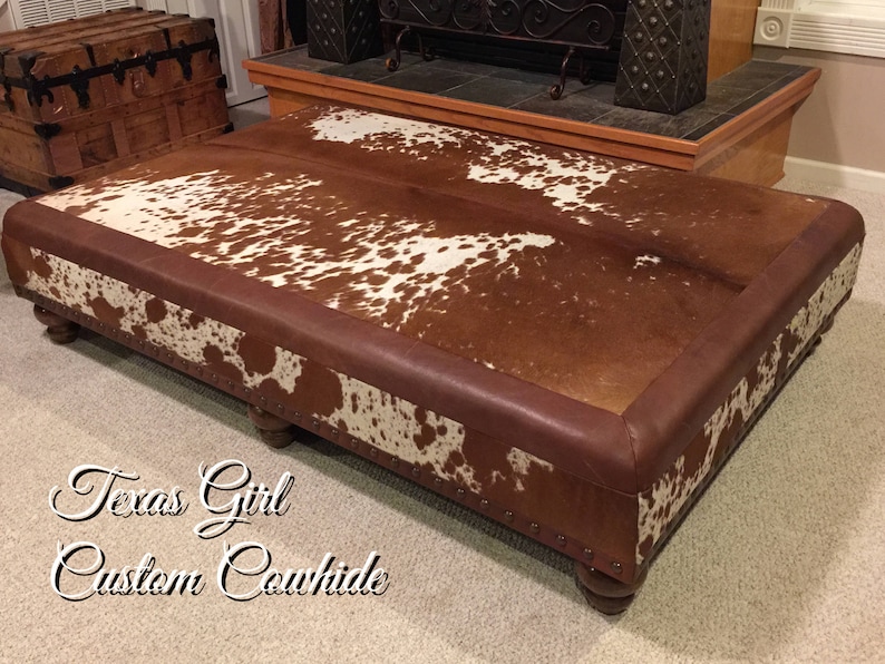 May include: A large, rectangular ottoman upholstered in brown leather with a cowhide top. The cowhide is brown and white with a spotted pattern. The ottoman has four wooden legs.