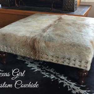 May include: A white and brown cowhide ottoman with a black and white cowhide rug underneath. The ottoman has a wooden frame and is decorated with metal studs.