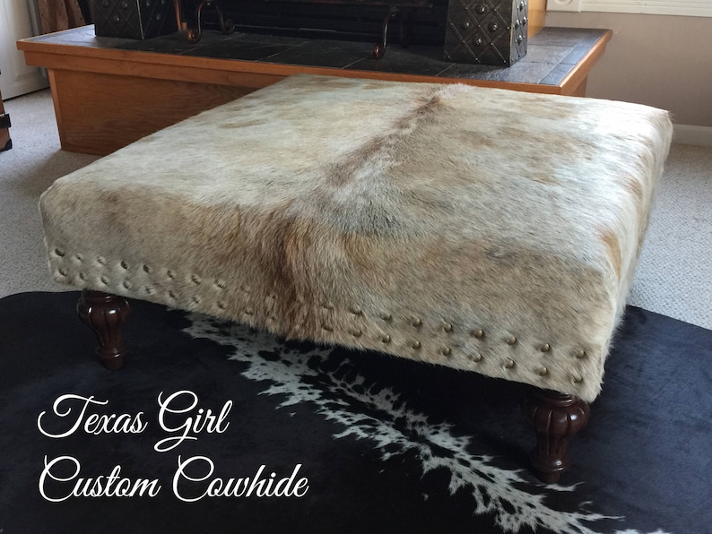 May include: A white and brown cowhide ottoman with a black and white cowhide rug underneath. The ottoman has a wooden frame and is decorated with metal studs.