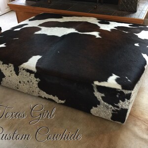 May include: A black and white cowhide ottoman with a brown patch. The ottoman is sitting on a white cowhide rug. The text "Texas Girl Custom Cowhide" is written on the rug.