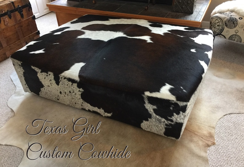 May include: A black and white cowhide ottoman with a brown patch. The ottoman is sitting on a white cowhide rug. The text "Texas Girl Custom Cowhide" is written on the rug.