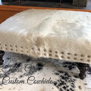 May include: A white cowhide ottoman with a tufted top and decorative nailhead trim. The ottoman has four dark wood legs and is sitting on a black and white cowhide rug.