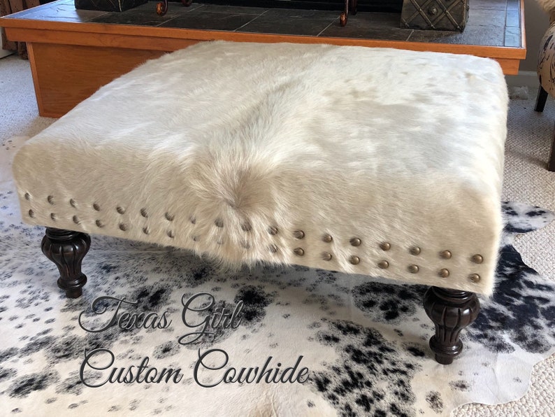 May include: A white cowhide ottoman with a tufted top and decorative nailhead trim. The ottoman has four dark wood legs and is sitting on a black and white cowhide rug.
