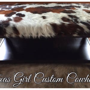 May include: A black wooden coffee table with a cowhide top and a lower shelf. The table has turned legs and is sitting on a cowhide rug.