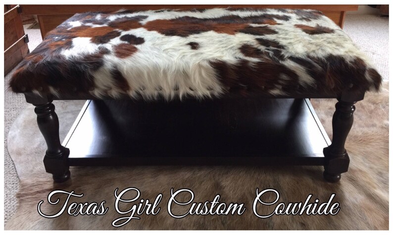 May include: A black wooden coffee table with a cowhide top and a lower shelf. The table has turned legs and is sitting on a cowhide rug.