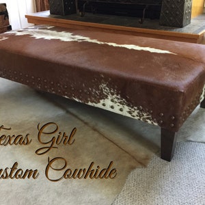 May include: A brown and white cowhide ottoman with dark brown wooden legs and metal studs around the edges. The ottoman is sitting on a light brown carpet. The text "Texas Girl Custom Cowhide" is written in cursive on the carpet.