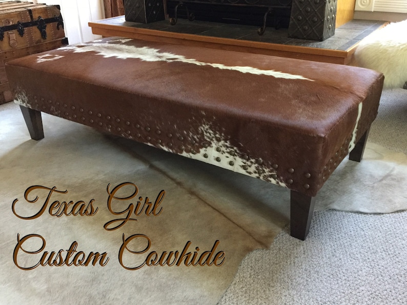 May include: A brown and white cowhide ottoman with dark brown wooden legs and metal studs around the edges. The ottoman is sitting on a light brown carpet. The text "Texas Girl Custom Cowhide" is written in cursive on the carpet.