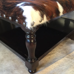 May include: A brown, black, and white cowhide ottoman with a dark wood base and turned legs. The ottoman has a decorative nailhead trim around the edge.