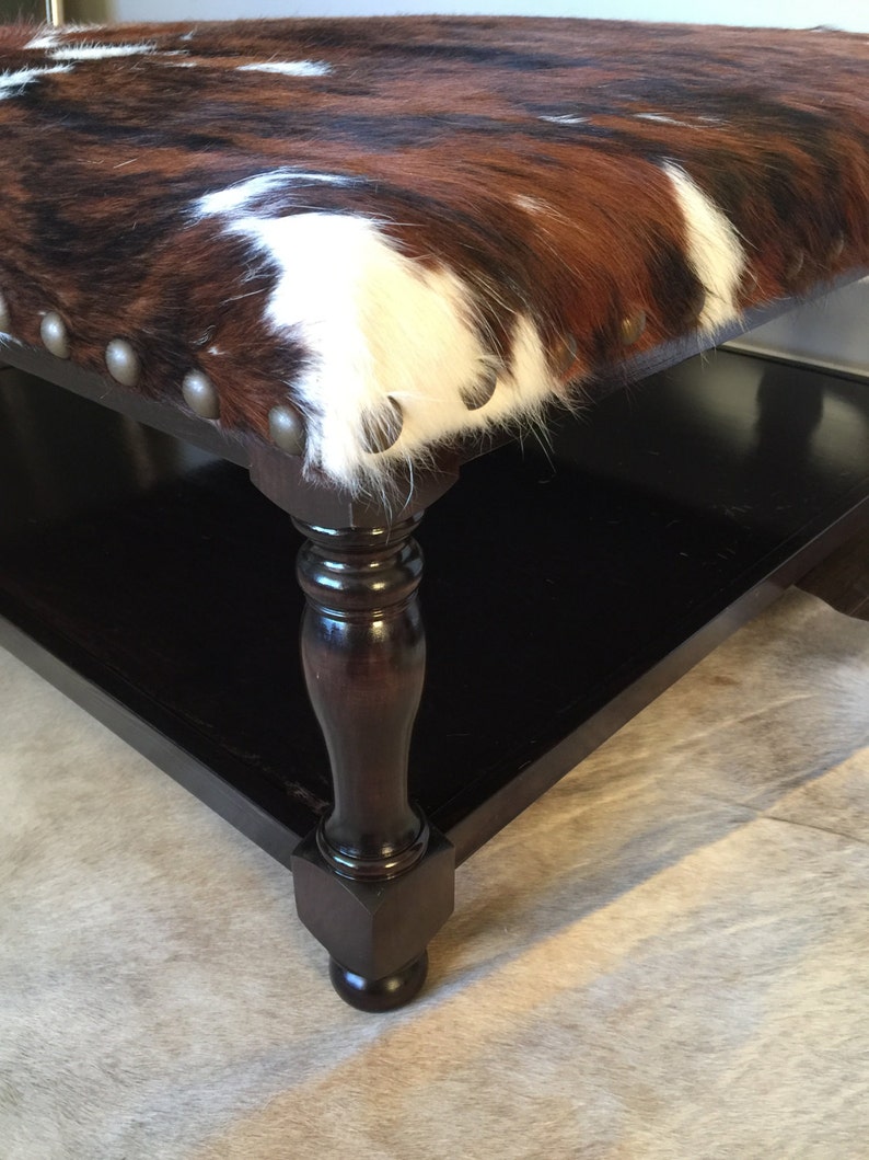 May include: A brown, black, and white cowhide ottoman with a dark wood base and turned legs. The ottoman has a decorative nailhead trim around the edge.