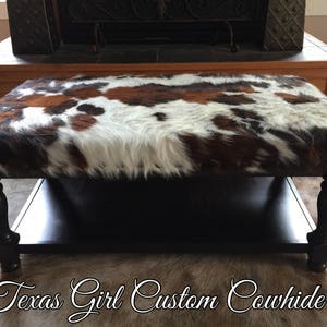 May include: A black wooden coffee table with a cowhide top. The table has a lower shelf and turned legs.