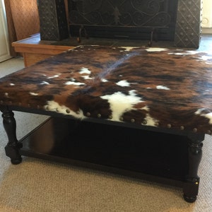 May include: A brown, white, and black cowhide ottoman with a black wooden base and a lower shelf. The ottoman has metal studs around the edge.