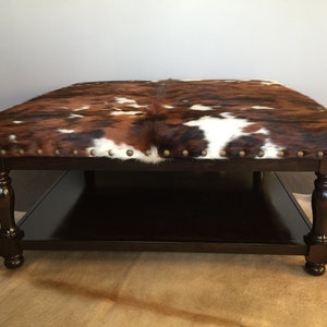 May include: A brown, black, and white cowhide ottoman with a dark wood frame and a lower shelf. The ottoman has a square shape and is decorated with brass tacks around the edges.