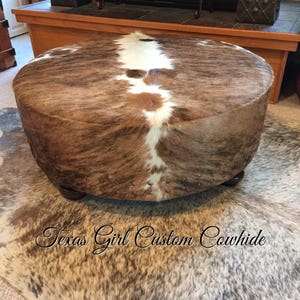May include: A round, brown and white cowhide ottoman with a brown leather base. The ottoman is sitting on a cowhide rug.