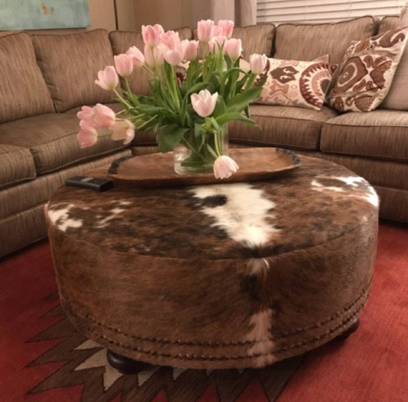 May include: A round brown and white cowhide ottoman with a wooden tray on top. The ottoman is decorated with a row of metal studs around the bottom edge. A bouquet of pink tulips is in a glass vase on the tray.