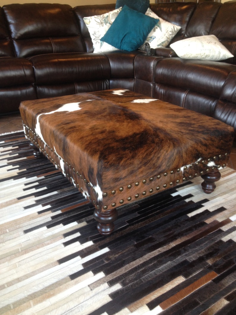 May include: A brown and white cowhide ottoman with studded trim sits on a black and white cowhide rug in front of a brown leather sectional sofa.